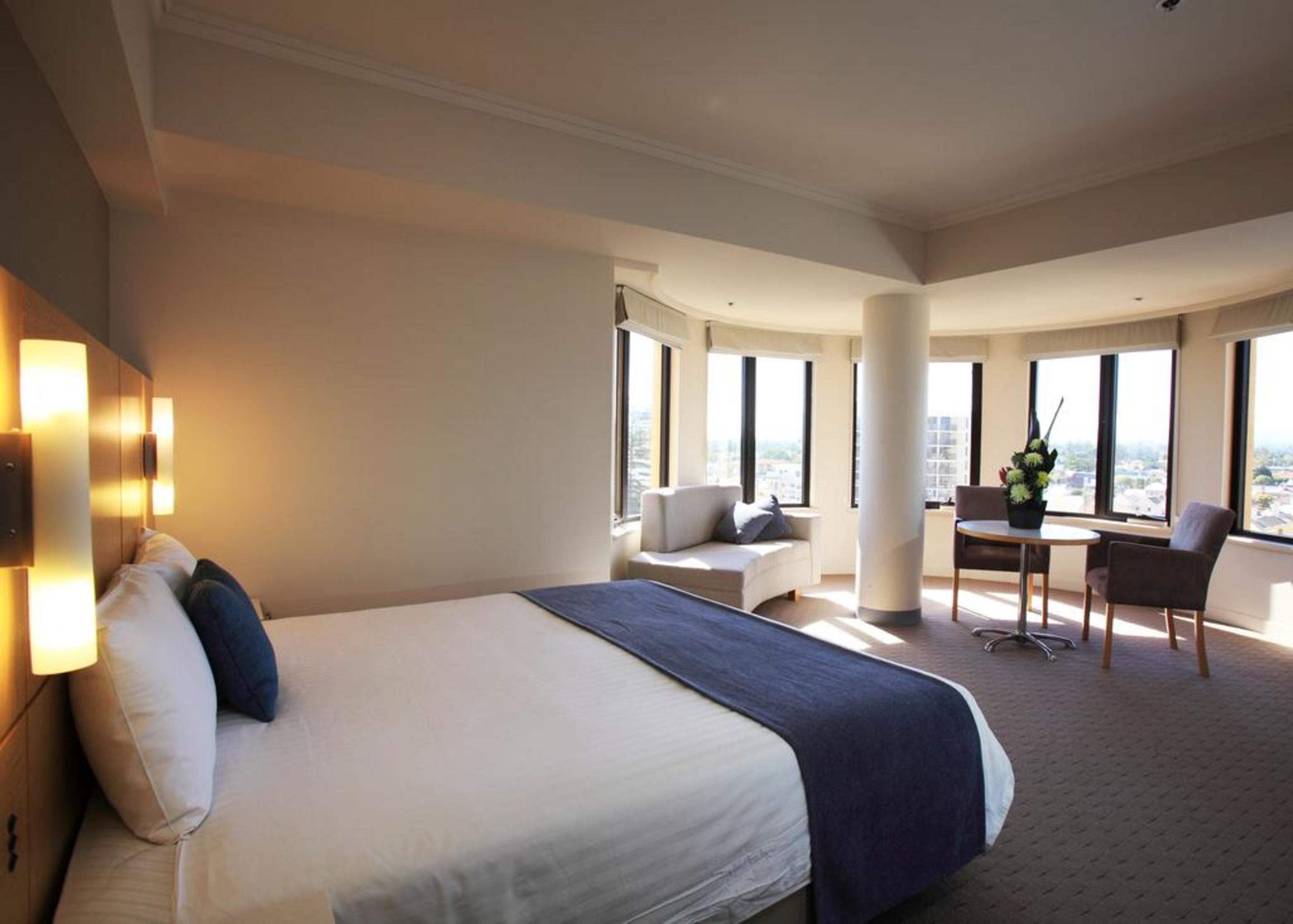 Stamford Grand Adelaide Hotel เกลเนลก์ ภายนอก รูปภาพ The photo shows a hotel room with a modern and spacious design. There is a large bed with white linens and a dark blue throw blanket. The room features multiple windows that allow plenty of natural light, and there is a small seating area near the wi