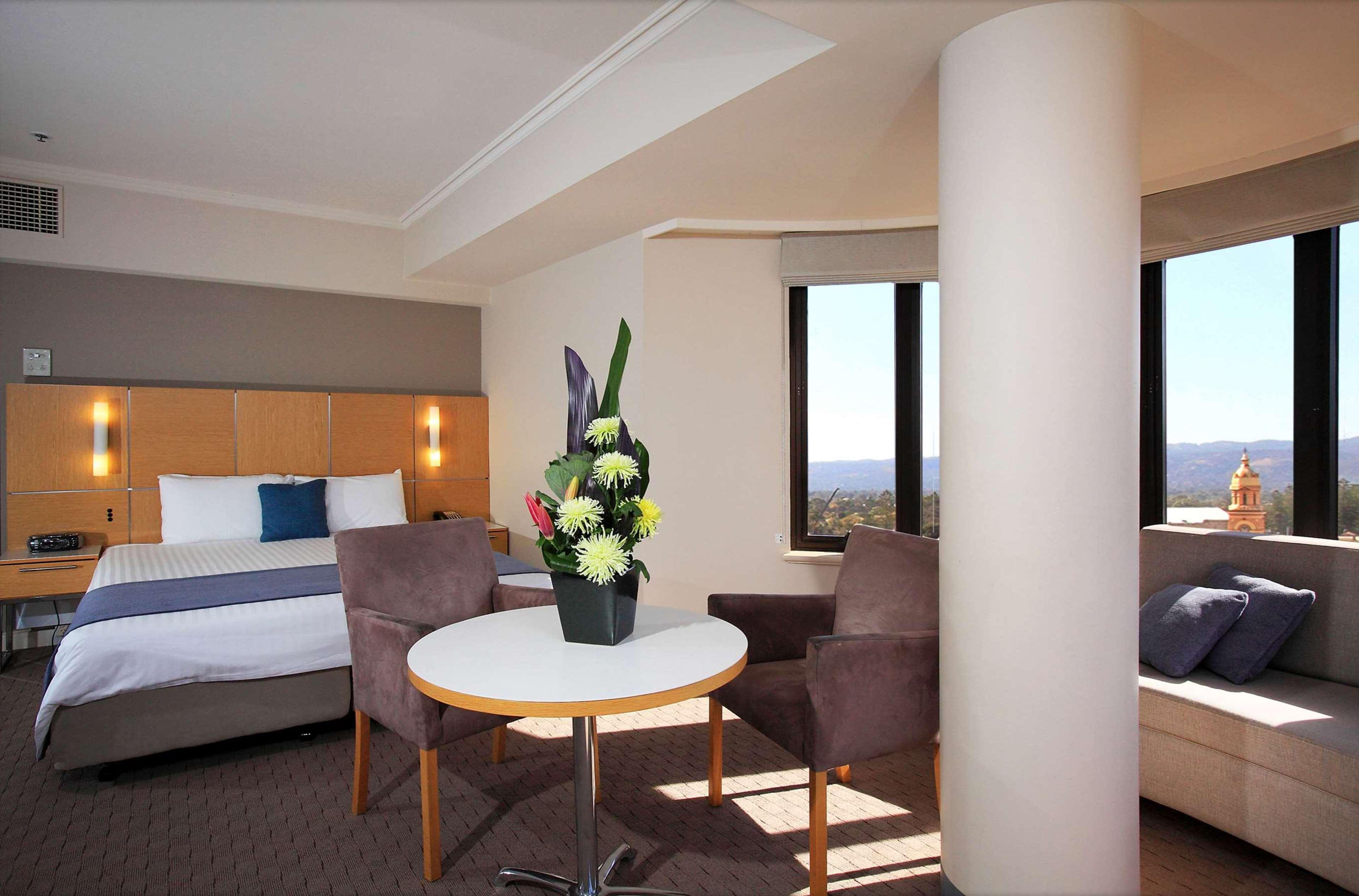Stamford Grand Adelaide Hotel เกลเนลก์ ภายนอก รูปภาพ The photo shows a hotel room with a modern design. On one side, there is a plush bed with white and blue bedding. In the center of the room, there's a small round table with two gray upholstered chairs around it. A vase of flowers sits on the table, 