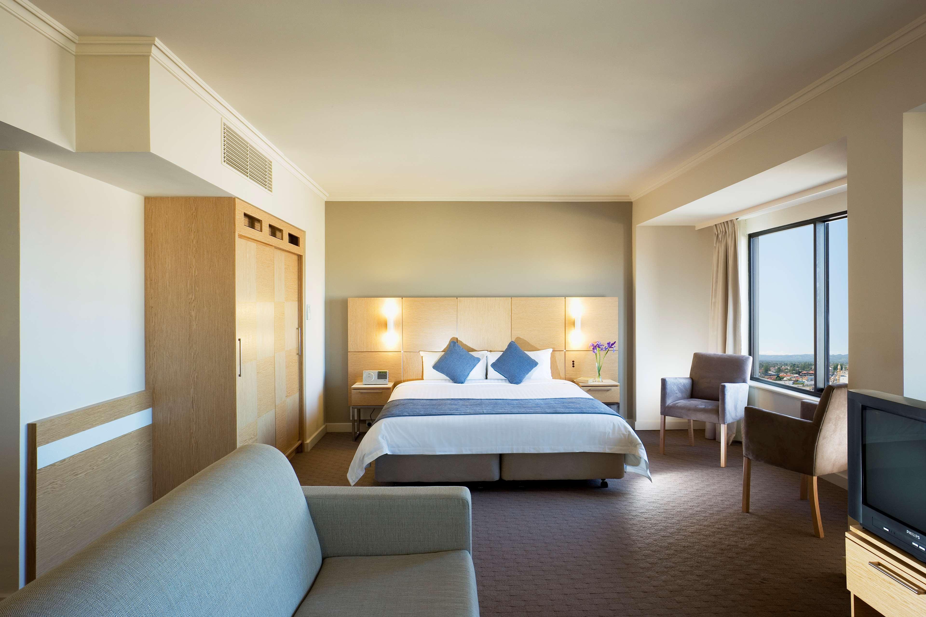 Stamford Grand Adelaide Hotel เกลเนลก์ ห้อง รูปภาพ The photo shows a modern hotel room featuring a large bed with two blue decorative pillows. There are bedside lamps on both sides of the headboard. To the left, there's a wooden wardrobe and a small table with a vase and a few amenities. On the right