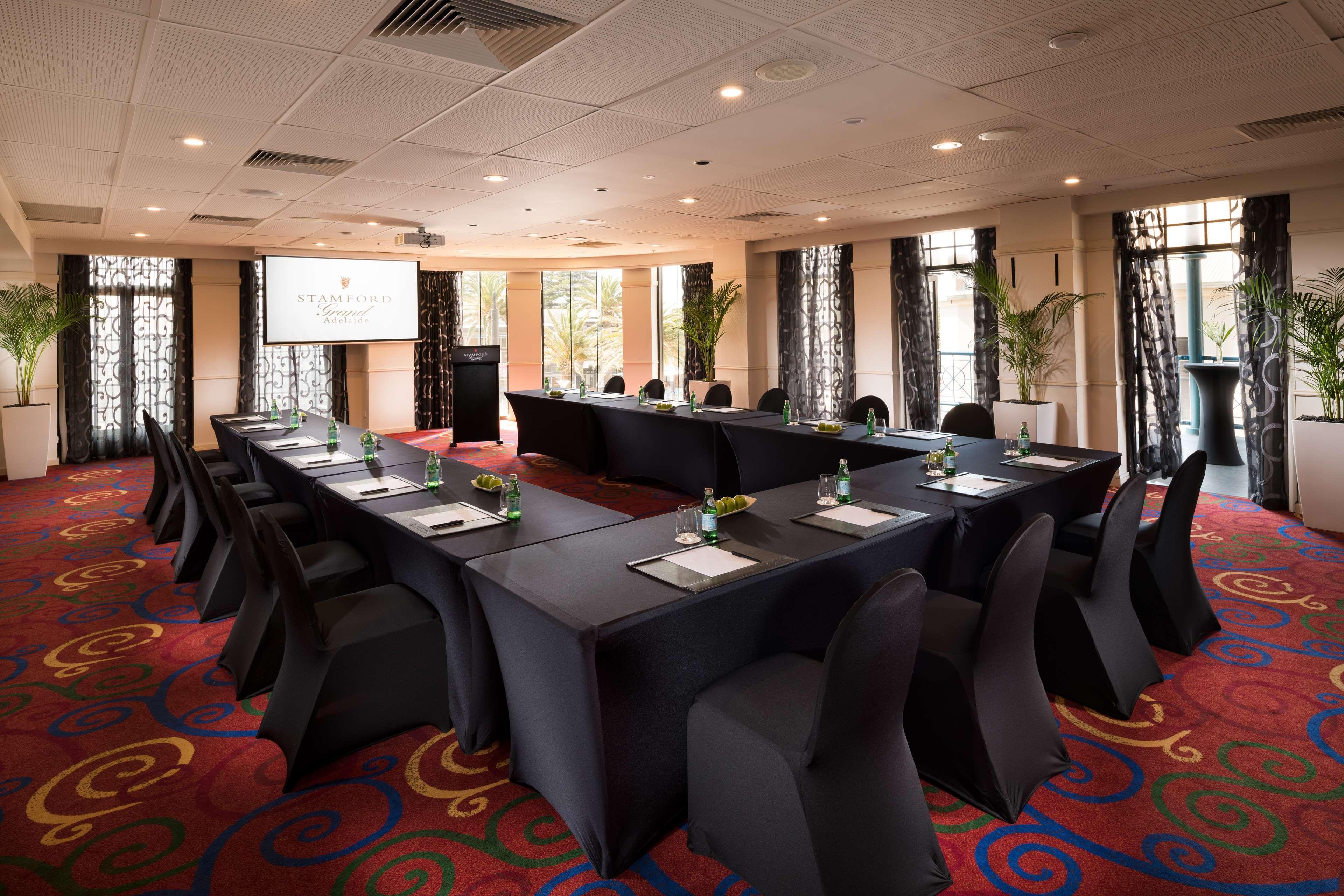 Stamford Grand Adelaide Hotel เกลเนลก์ ภายนอก รูปภาพ The photo shows a conference room set up for a meeting or presentation. There are several tables arranged in a U-shape, covered with black tablecloths. Each table has a notepad and a small bottle of water on it. The chairs around the tables are also 