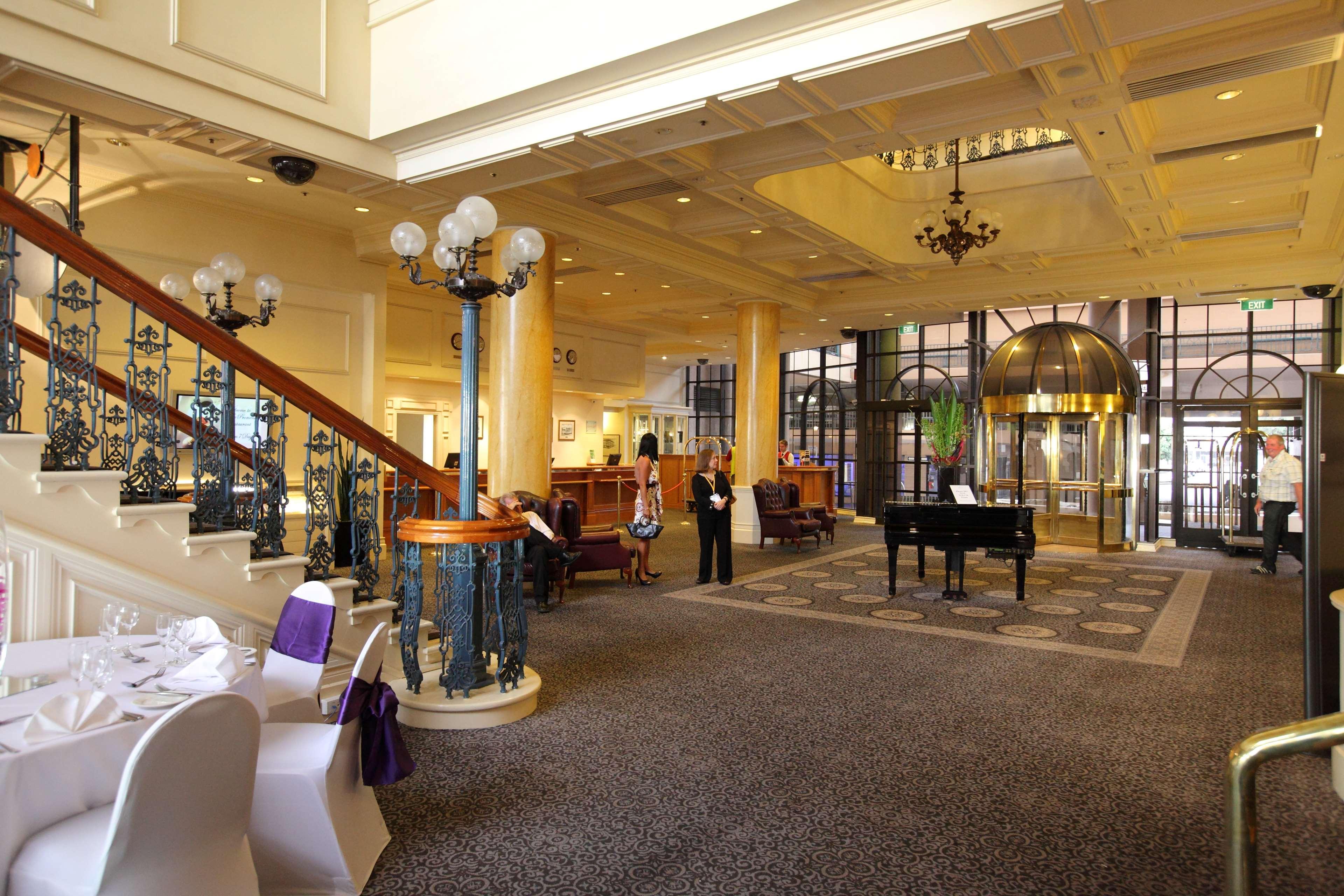 Stamford Grand Adelaide Hotel เกลเนลก์ ภายใน รูปภาพ The photo shows a luxurious interior of a hotel or a restaurant lobby. There is an elegant staircase with ornate railings leading to an upper level. The space features high ceilings with decorative chandeliers and ample natural light coming through l