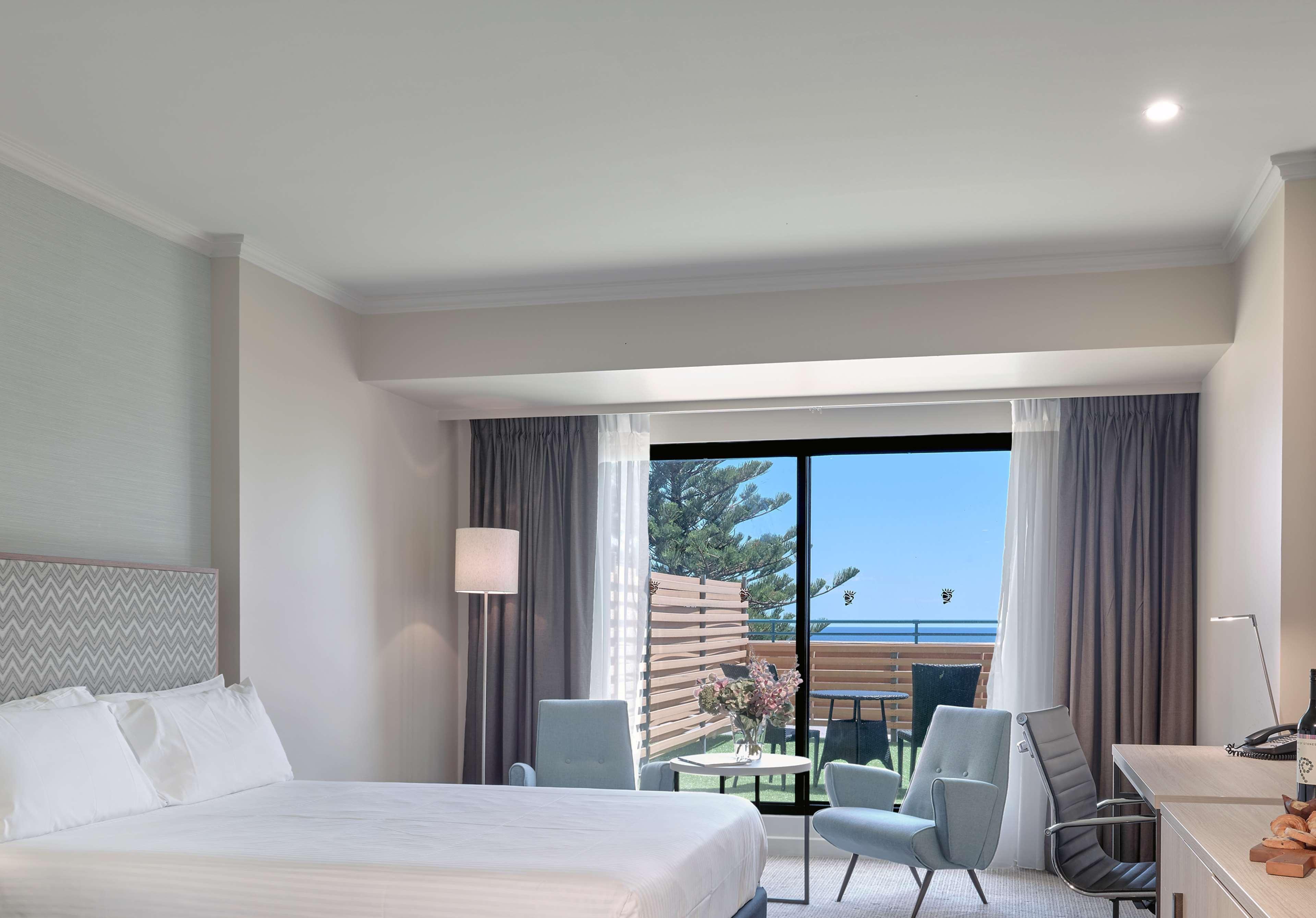 Stamford Grand Adelaide Hotel เกลเนลก์ ภายนอก รูปภาพ The photo shows a modern hotel room featuring a large bed with white bedding. There are two armchairs positioned near a window that offers a view of an outdoor area, possibly a balcony or patio with greenery. The room has light-colored walls and curt