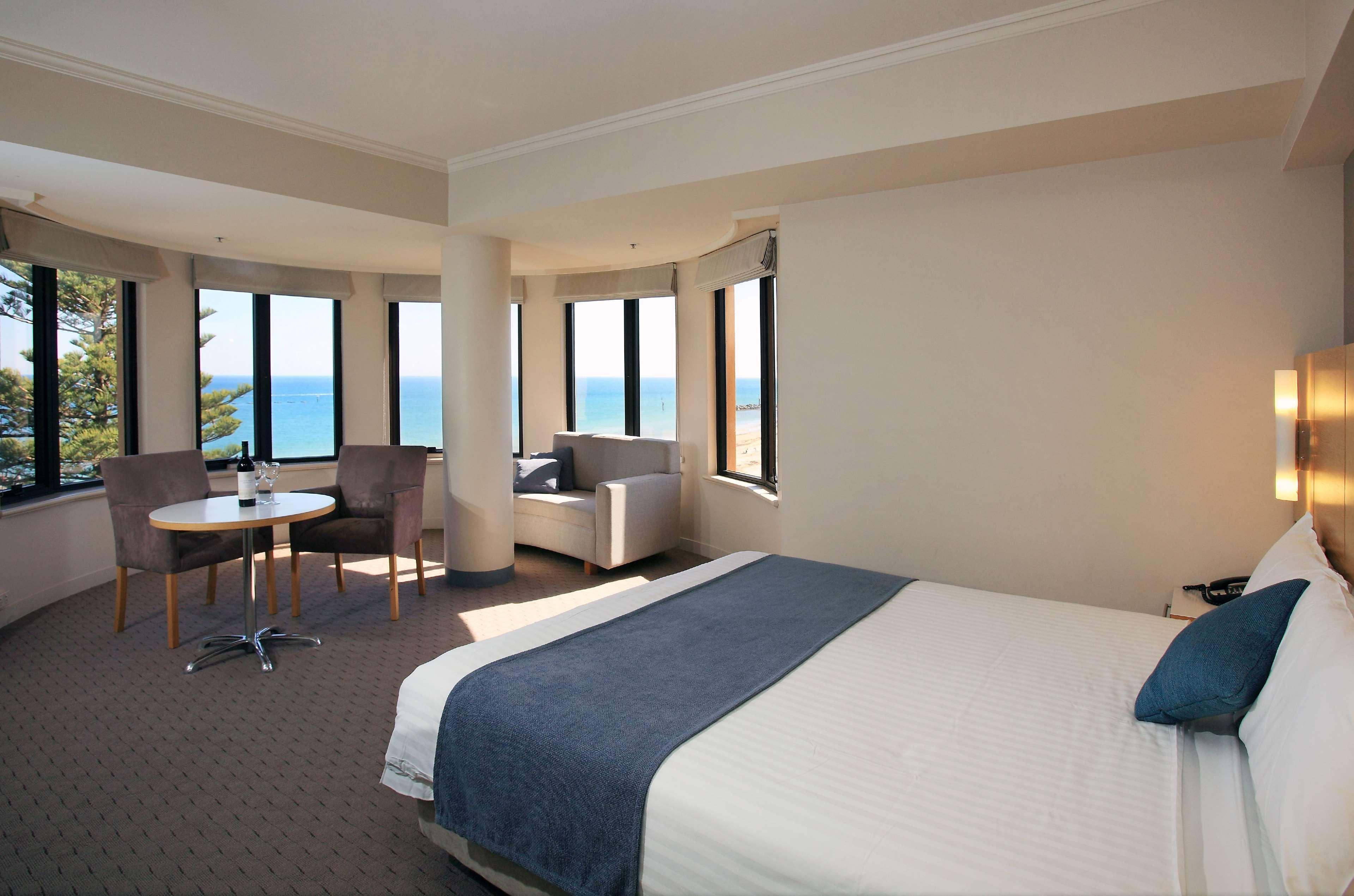 Stamford Grand Adelaide Hotel เกลเนลก์ ภายนอก รูปภาพ The photo shows a well-lit hotel room featuring large windows that offer a view of the ocean. The room is arranged with a comfortable bed covered in white linens and a navy blue throw blanket. There is a small table and chairs set up beside the windo