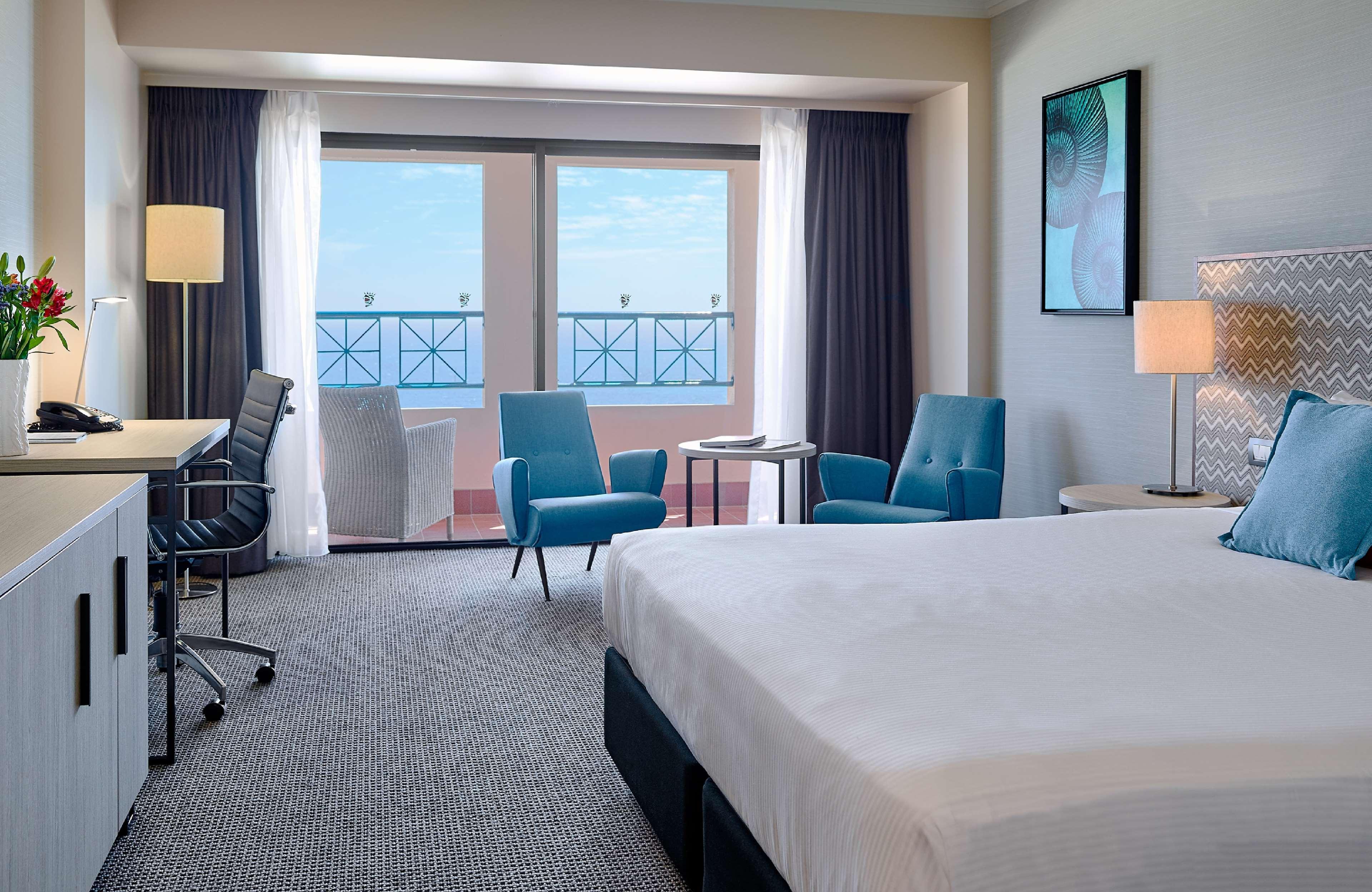 Stamford Grand Adelaide Hotel เกลเนลก์ ภายนอก รูปภาพ The image shows a hotel room featuring a large bed with a white duvet. There are two bright blue chairs and a small table near a large window that provides a view outside, possibly to a balcony. The room includes a workspace with a desk and an office
