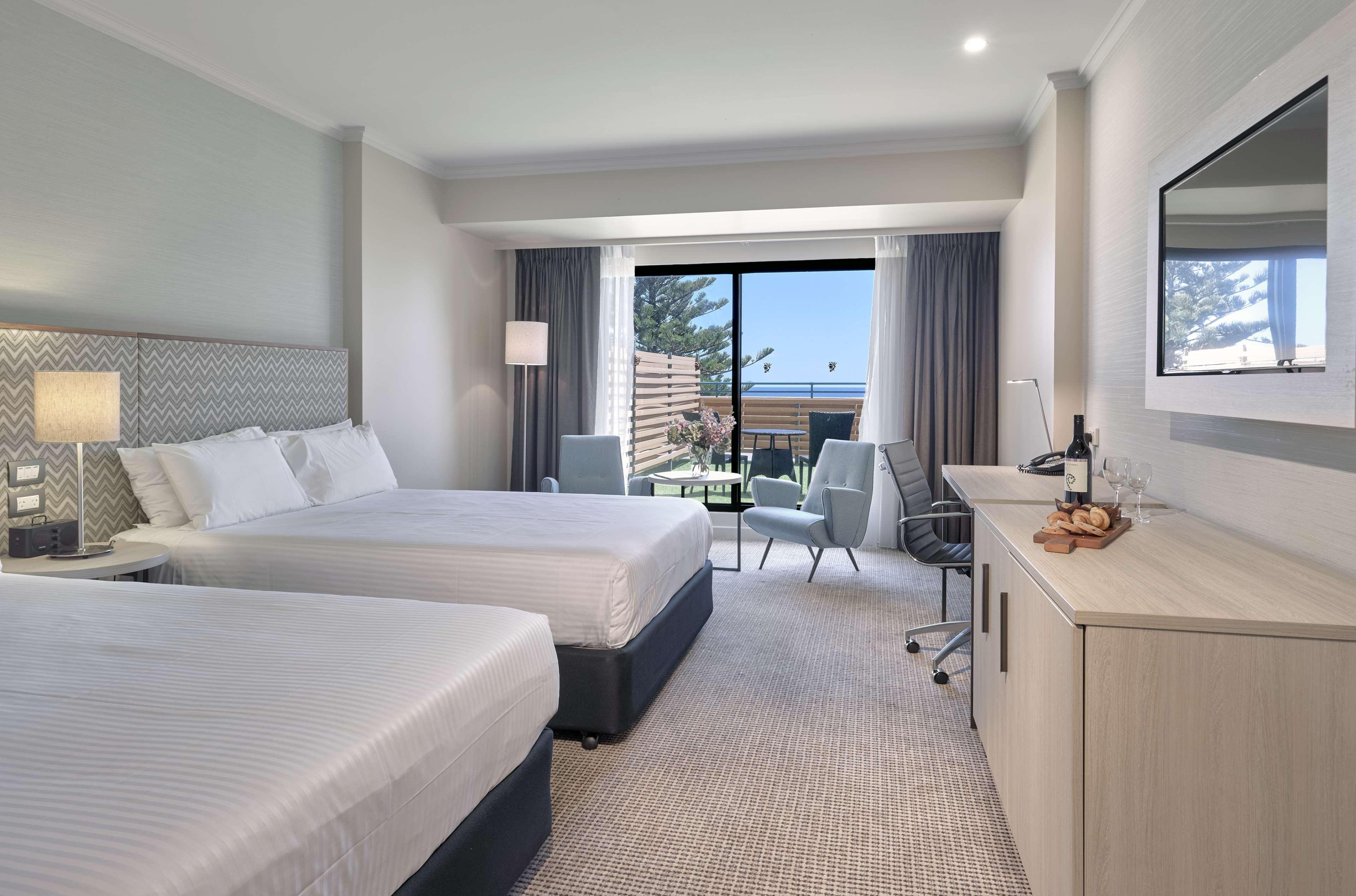 Stamford Grand Adelaide Hotel เกลเนลก์ ภายนอก รูปภาพ The photo shows a modern hotel room featuring two beds with white linens. There's a large window that provides natural light and a view of a balcony area. The room includes two chairs and a small table, creating a cozy sitting area. A television is m
