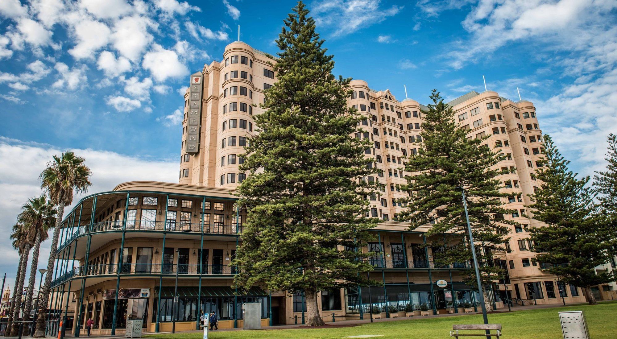 Stamford Grand Adelaide Hotel เกลเนลก์ ภายนอก รูปภาพ The image shows a large building with a distinctive architectural style, characterized by a tan or beige color. The structure appears to have multiple floors and rounded edges. In front of the building, there are several tall green trees, which add t