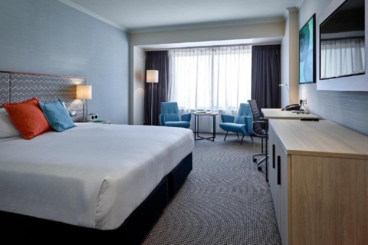 Stamford Grand Adelaide Hotel เกลเนลก์ ภายนอก รูปภาพ The photo shows a modern hotel room with a clean and minimalist design. In the center, there is a large, neatly made bed with white linens and two decorative pillows, one orange and one blue. To the right, there is a small wooden cabinet with a telev